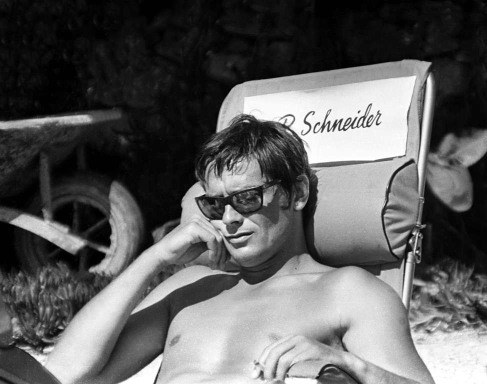 BRADEL104 - 1672511 Affiche Delon La Piscine : photographie d'art en noir et blanc d'Alain Delon torse nu avec des lunettes de soleil, assis sur une chaise de tournage au nom de Romy Schneider en 1968.