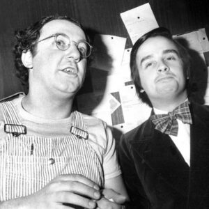 Affiche Coluche Le Luron : photographie d'art en noir et blanc de 1975 montrant Coluche en salopette à rayures et Thierry Le Luron en smoking avec un nœud papillon, capturés en coulisses.
