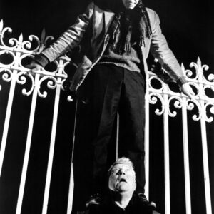 Affiche Singe en Hiver : photographie en noir et blanc de 1962 montrant Jean-Paul Belmondo debout sur les épaules de Jean Gabin, escaladant une imposante grille en fer forgé la nuit.
