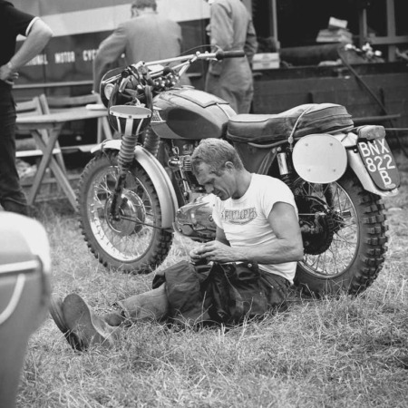 Affiche Steve McQueen Triumph : photographie vintage en noir et blanc de 1964 montrant Steve McQueen assis dans l'herbe, concentré, à côté de sa moto Triumph de compétition lors des Six Jours Internationaux d'Enduro.