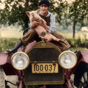 Affiche cochon vintage :Photographie d'époque colorisée de 1916 montrant une jeune femme en salopette assise sur le capot d'une voiture ancienne Stutz à New York, tenant un petit cochon.